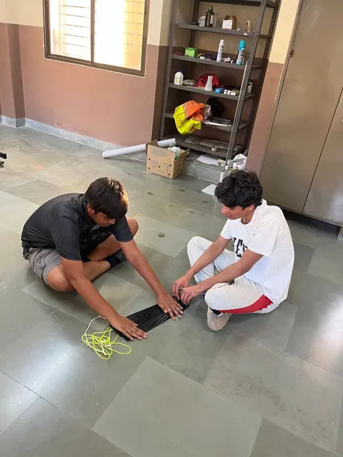 Two students sit on a tiled floor making a parachute, with a shelf of equipment behind them.