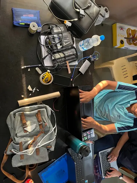Overhead view of a crowded worktable with tools, a laptop, bags and rocket parts.