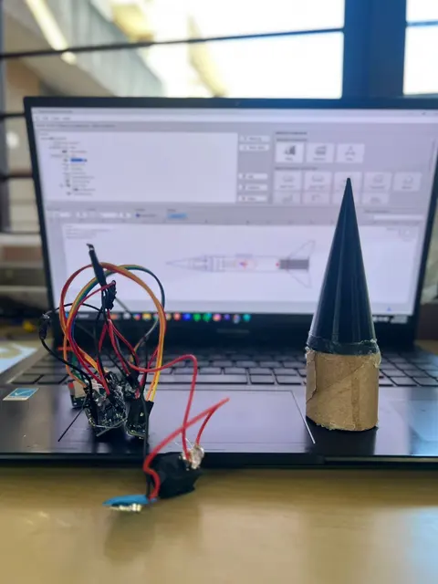 A breadboard with a microcontroller and wires beside a black nose cone, in front of a laptop.