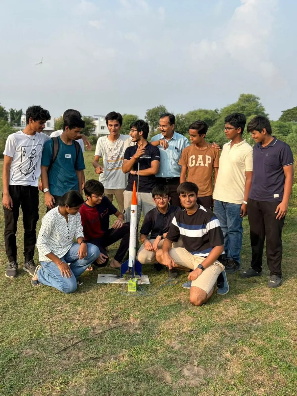 The team poses in a grassy field around SRM, white with an orange nose and blue fins, on its launch board.