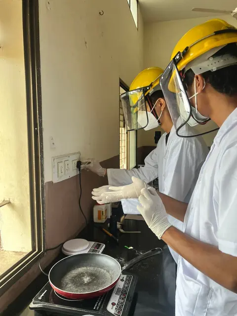 Two team members in hard hats with face shields, lab coats and gloves work at a stove by an open window.