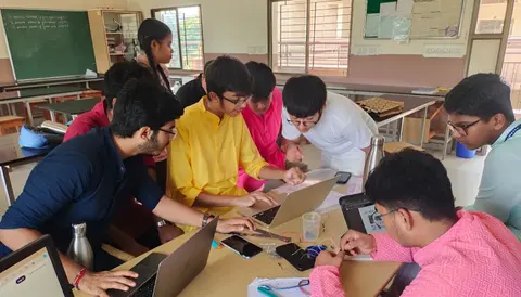 A group of students gathered around laptops on a classroom table.